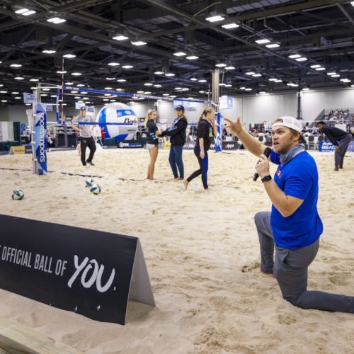 Man with a microphone kneeling in the sand of a volleyball court in a convention center